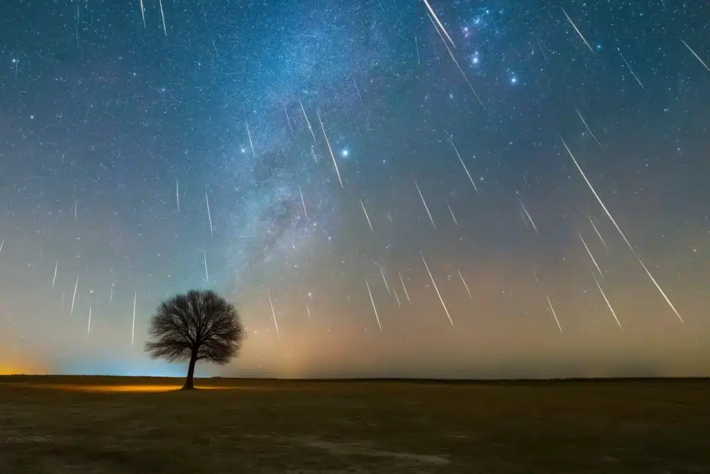 Geminid Meteor Shower 2025 streaks of bright meteors falling across a clear night sky above a lone tree in an open field.
