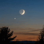 A waxing crescent moon and the planet Saturn appearing close together during the Moon and Saturn Conjunction in the evening sky over silhouetted trees.